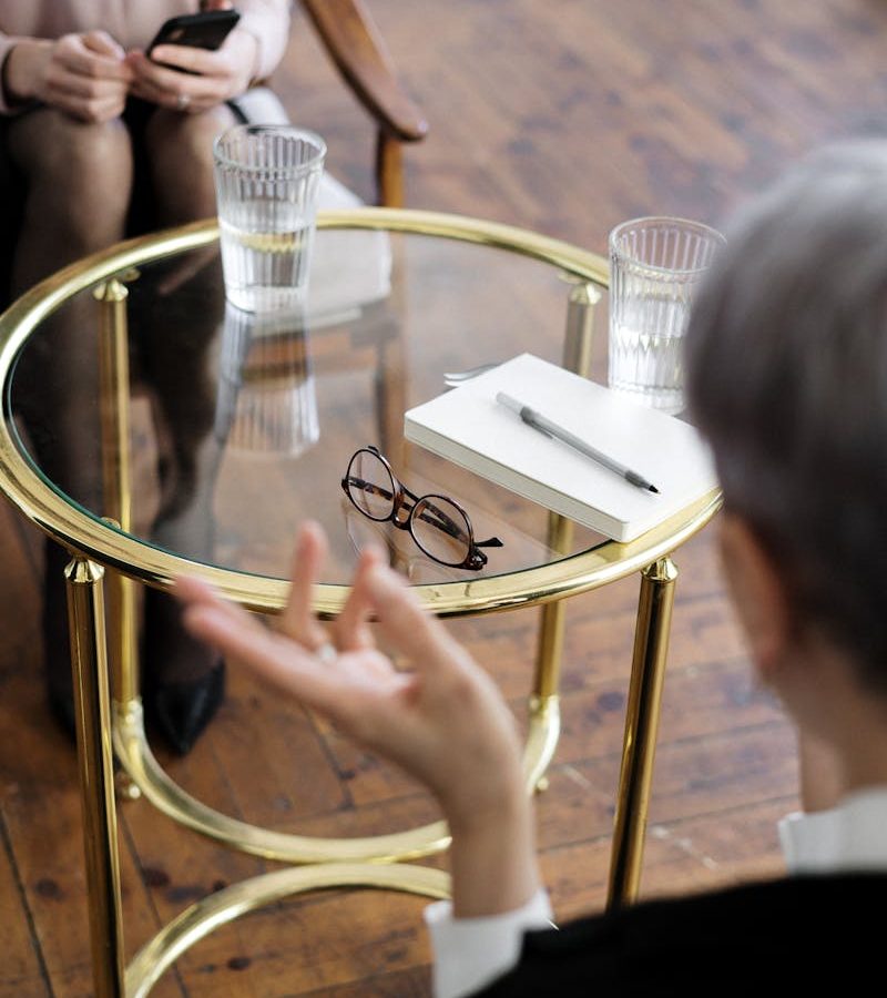 Two adults engaged in a therapy session, seated at a glass table with notebooks and water glasses.